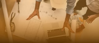 Foto de cima de duas pessoas trabalhando em uma mesa, nela há algumas papéis com desenhos técnicos e em cima um laptop